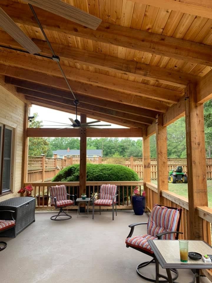 Covered wooden porch with chairs, tables, and a ceiling fan, overlooking a backyard with green bushes and lawn.
