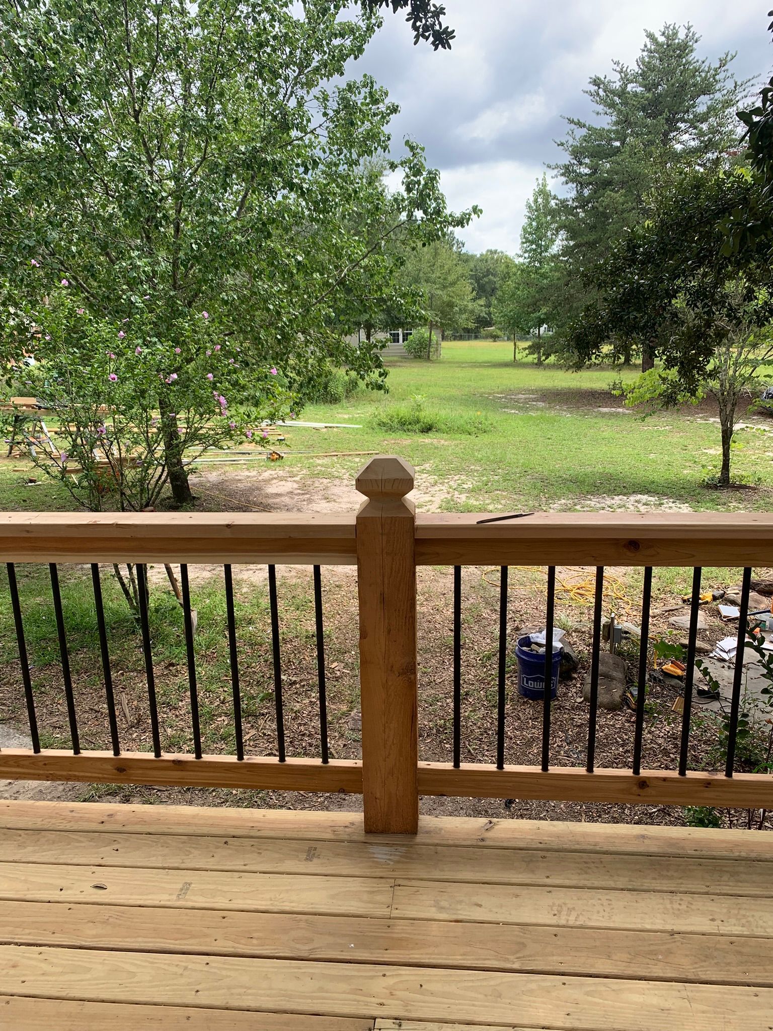 Wooden deck with railing overlooking a green field, trees, and a cloudy sky.