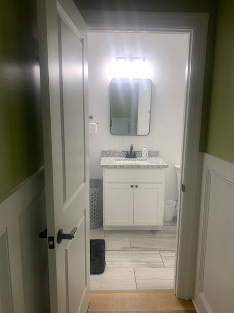 Open white door leads to a small bathroom with white vanity, mirror, and light fixture. Green walls.