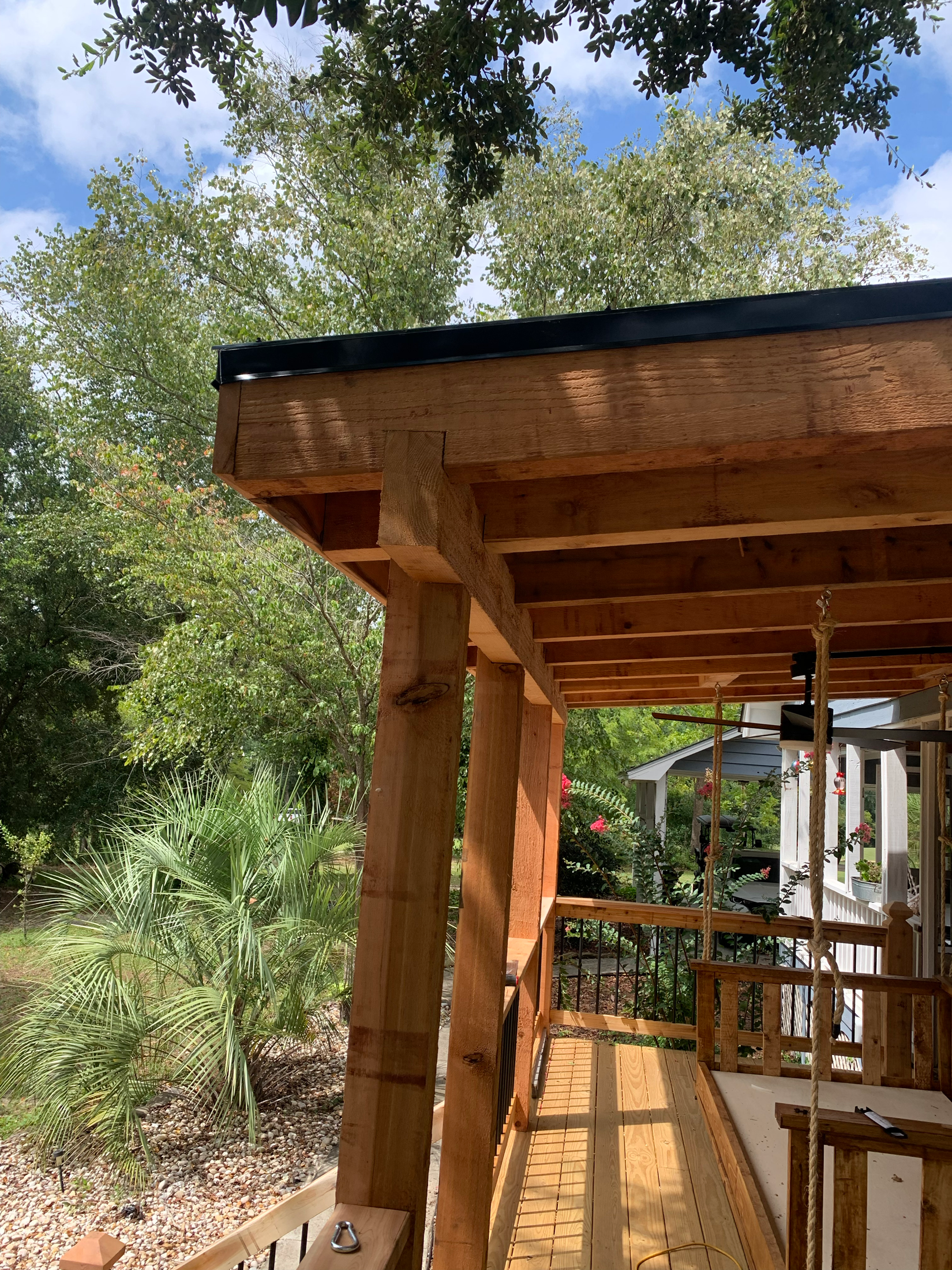 Wooden porch with a roof, trees, and a house in the background on a sunny day.