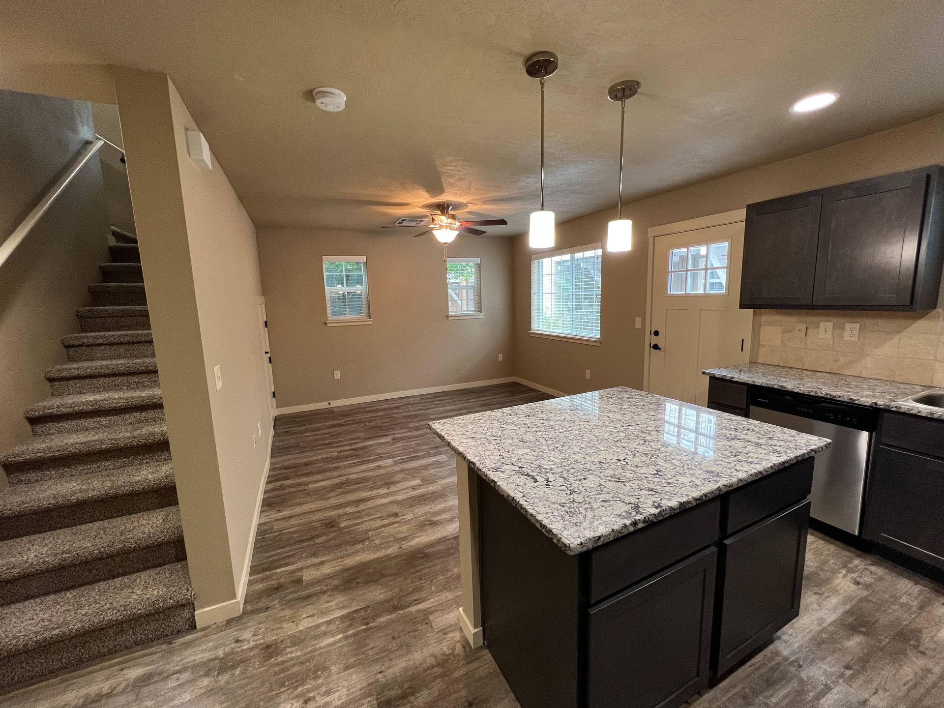 Interior view of a kitchen and living area with a staircase. Dark cabinets, granite countertop, and wood flooring.
