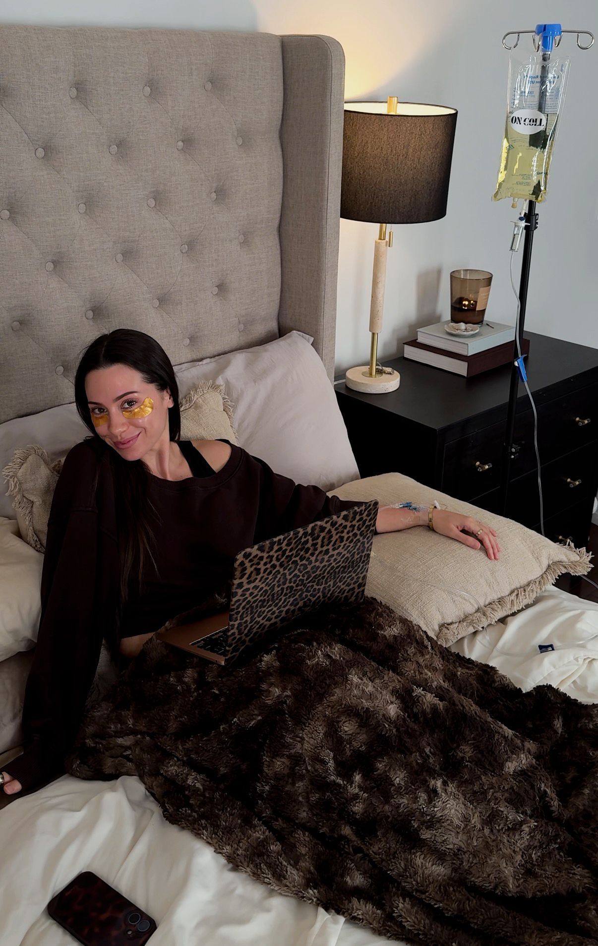 Woman reclining in bed with a laptop, under a dark patterned blanket beside a lamp and IV stand.
