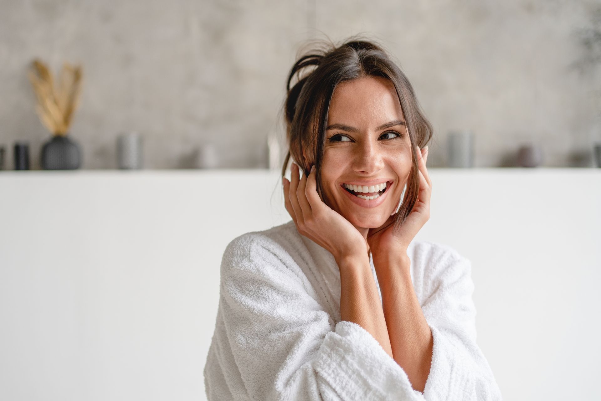 Smiling person in a white robe with hands on cheeks, standing indoors against a light wall.