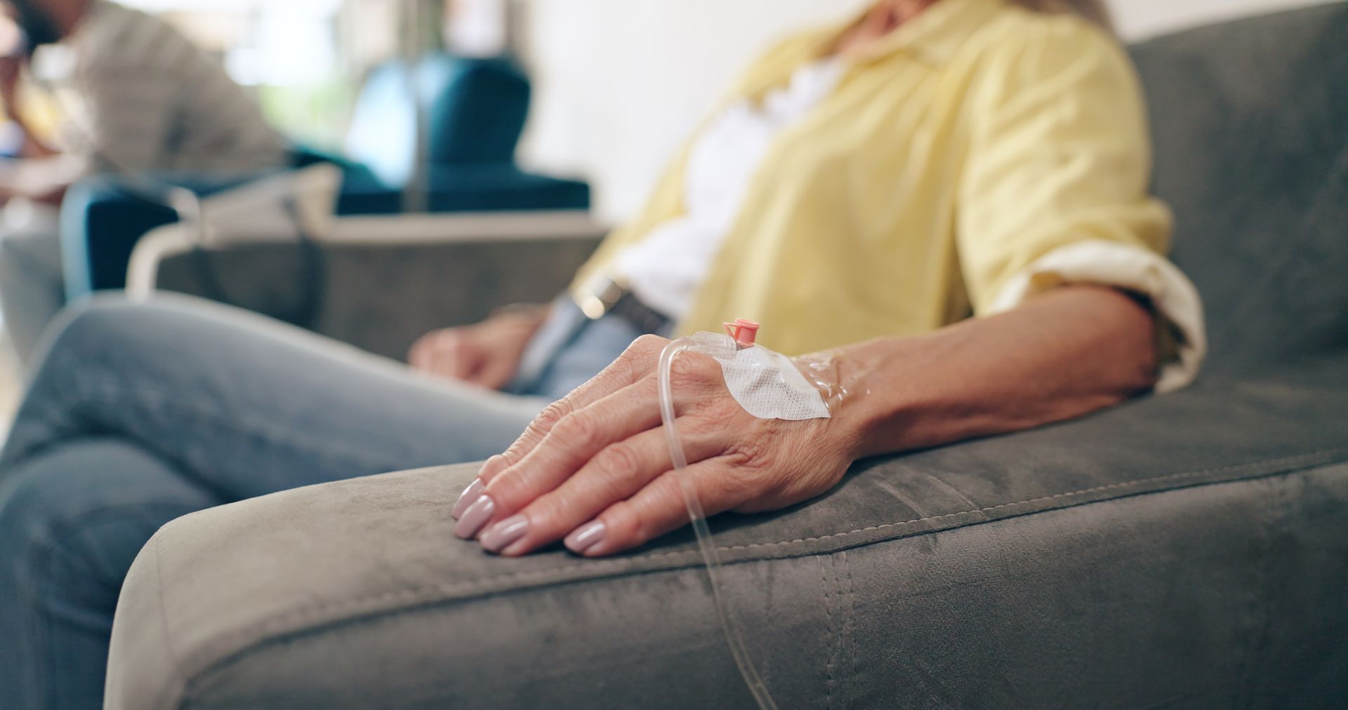Person resting on a couch with an IV in their hand, wearing a yellow shirt and gray pants.