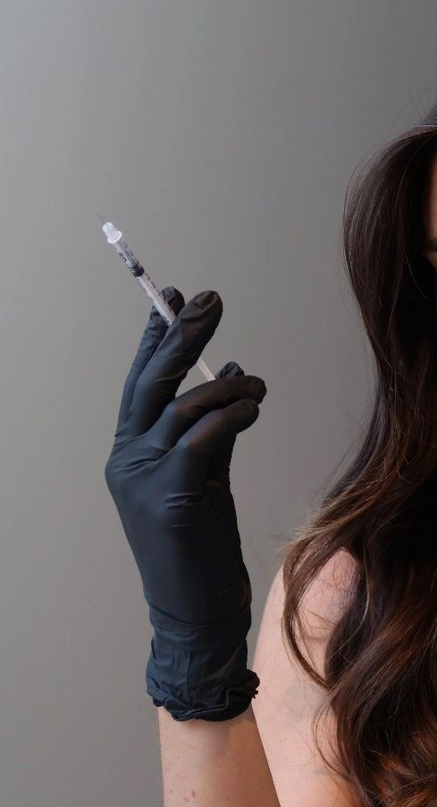 Gloved hand holding a syringe beside a person’s bare shoulder against a plain background