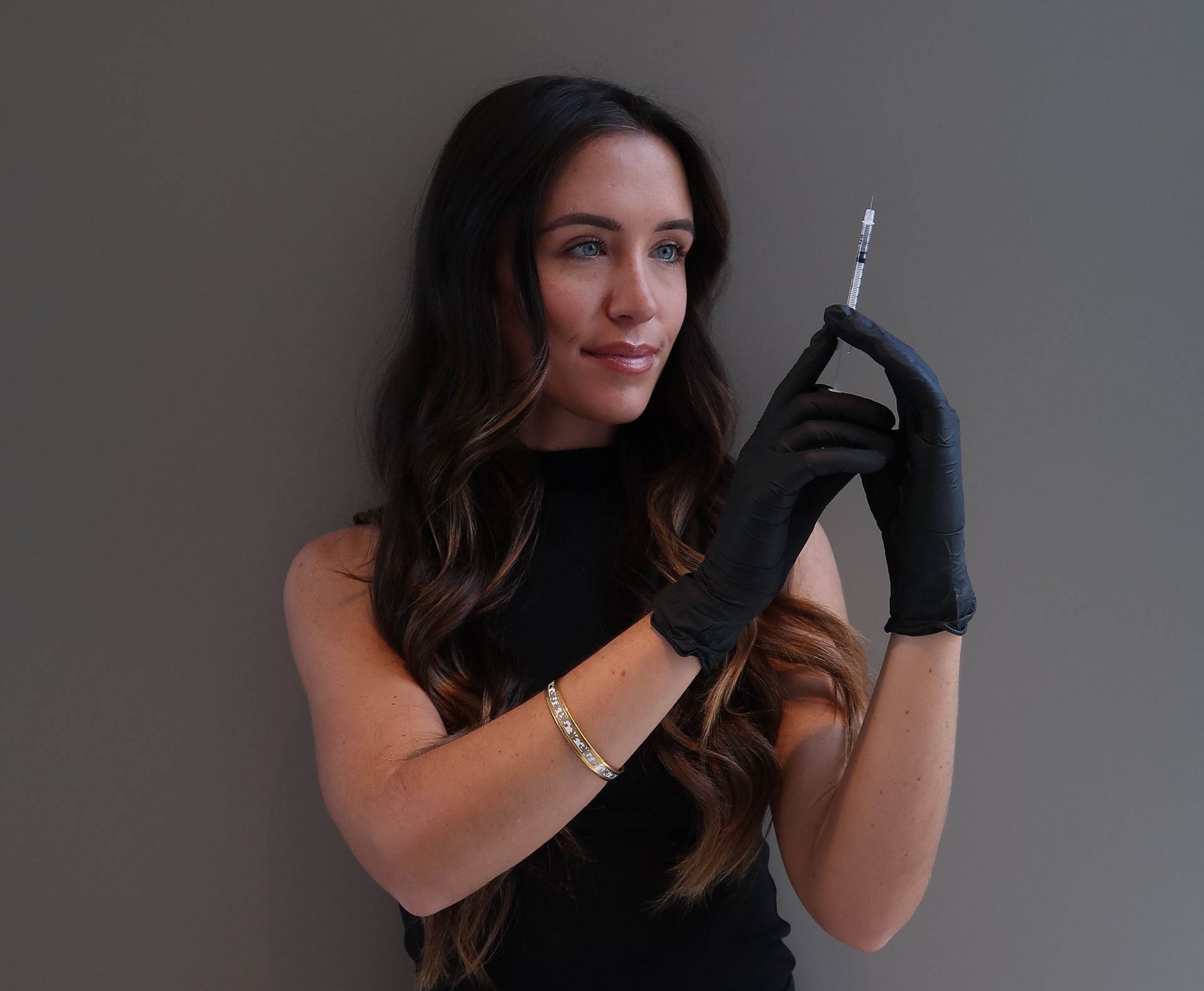 Woman in black gloves holding a syringe, standing against a gray background.