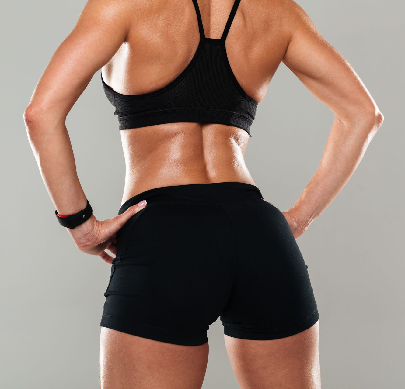 Back view of a fit athlete in black sportswear, standing with hands on hips against a gray background