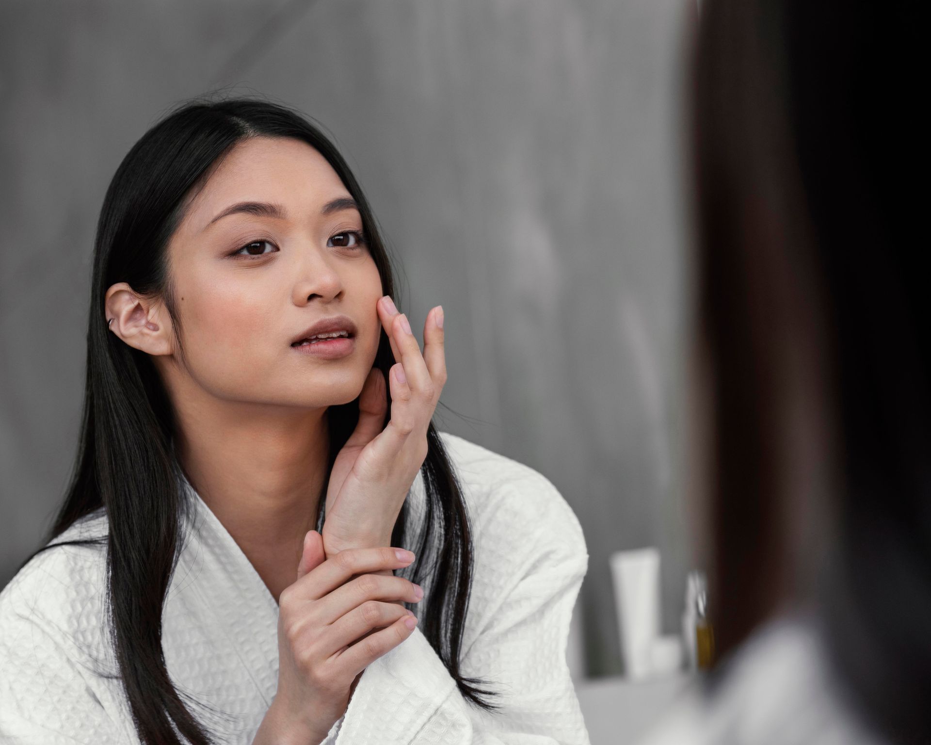 Woman in white robe applying skincare to her cheek while looking in a mirror