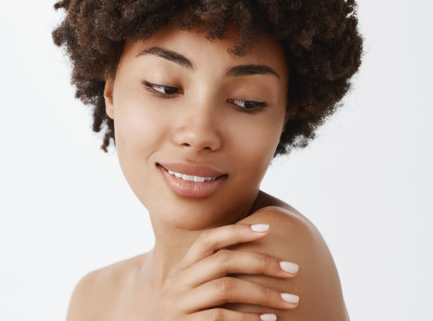 Smiling person with curly hair looking down, resting a hand on their shoulder against a white background