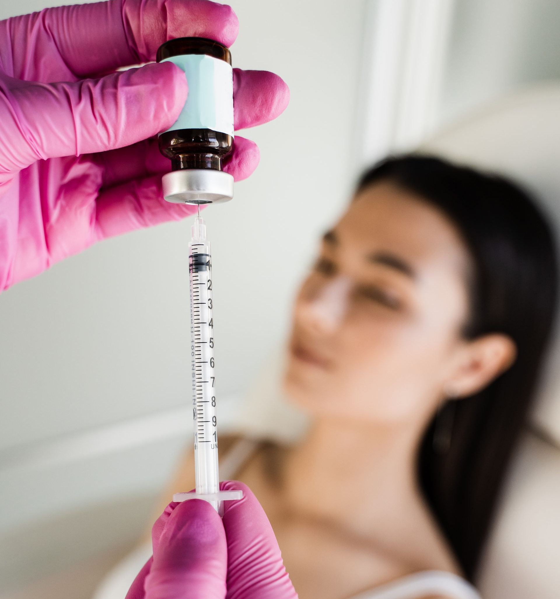 Gloved hands holding a syringe and vial near a blurred patient in a clinical setting