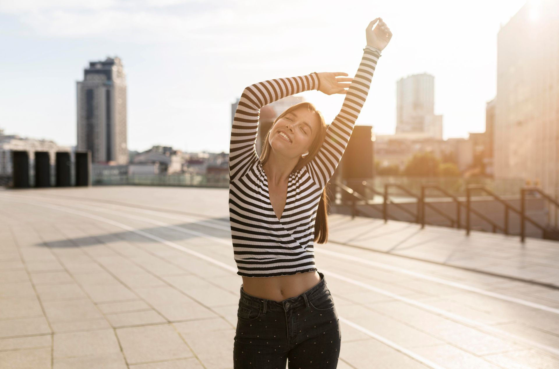 Person stretching with arms raised on a sunny rooftop with city skyline in the background