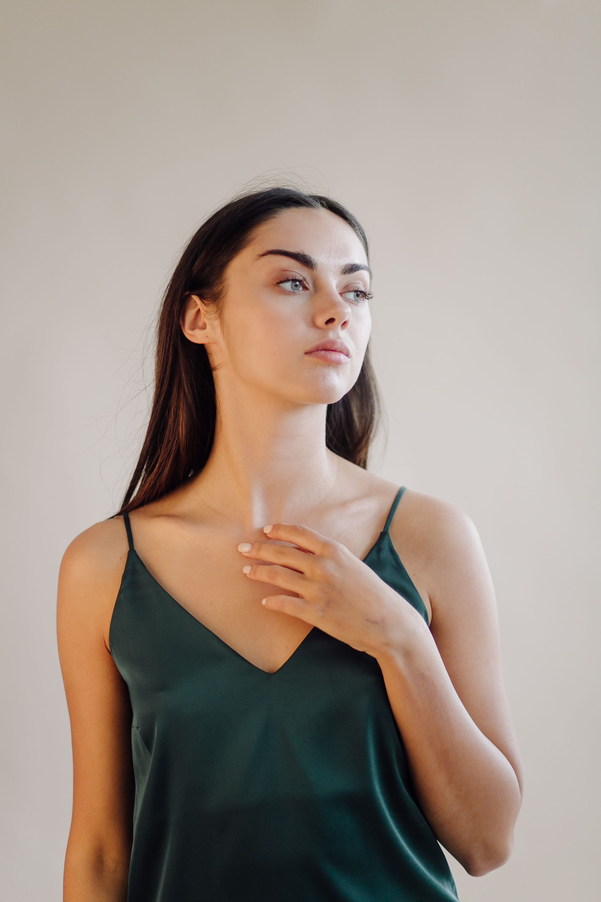 Woman in a dark green camisole, looking upward with one hand on her chest against a plain beige background