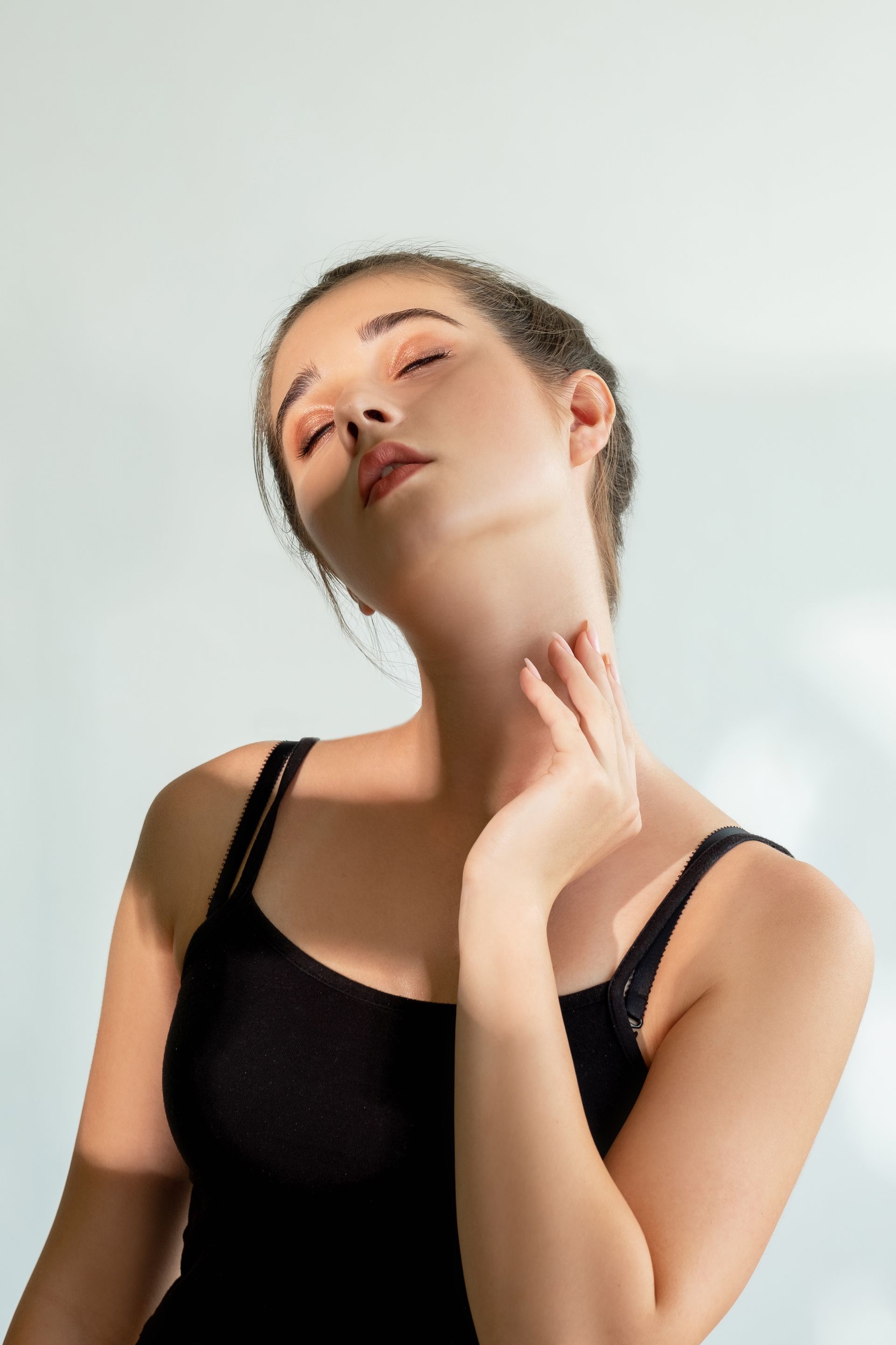 Woman in a black tank top posing with hand on neck against a light background