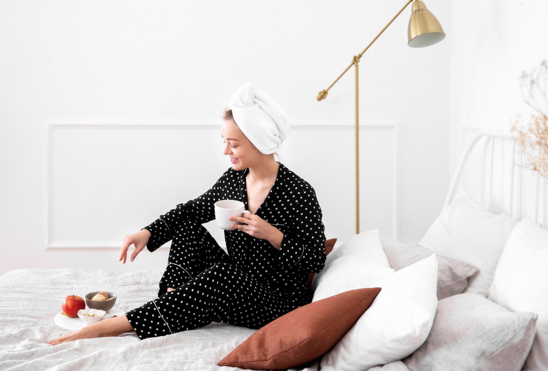 Woman in a white towel sits on a bed with a teacup, beside pillows, fruit, and a gold floor lamp