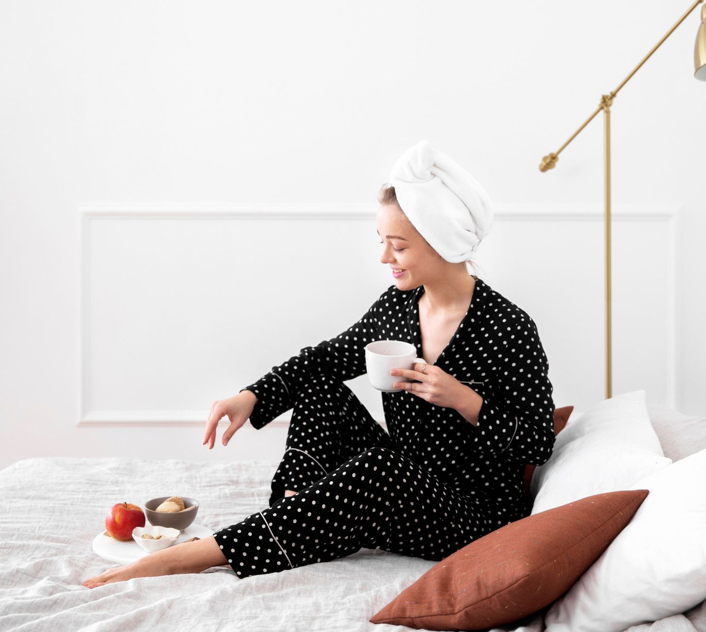 Woman in black polka-dot pajamas sits on a bed, holding a mug and relaxing in a bright bedroom.