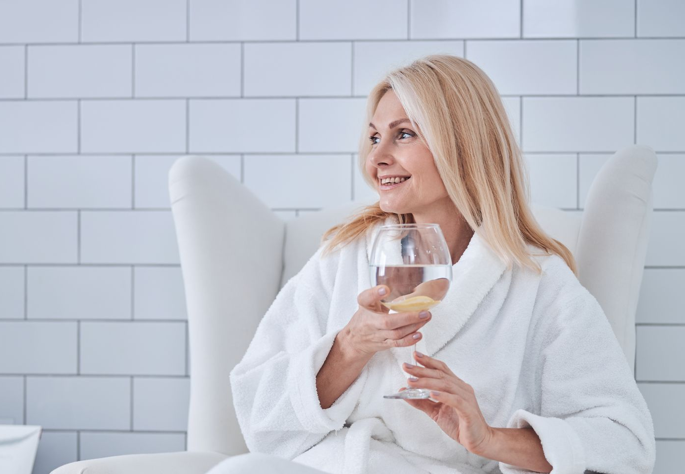 Woman in a white robe smiling and holding a glass of water in a bright tiled room