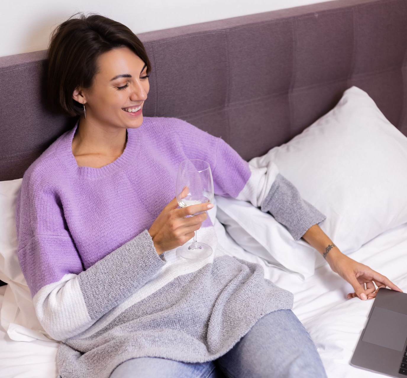 Woman smiling on a bed, holding a glass of water and looking at a laptop nearby.