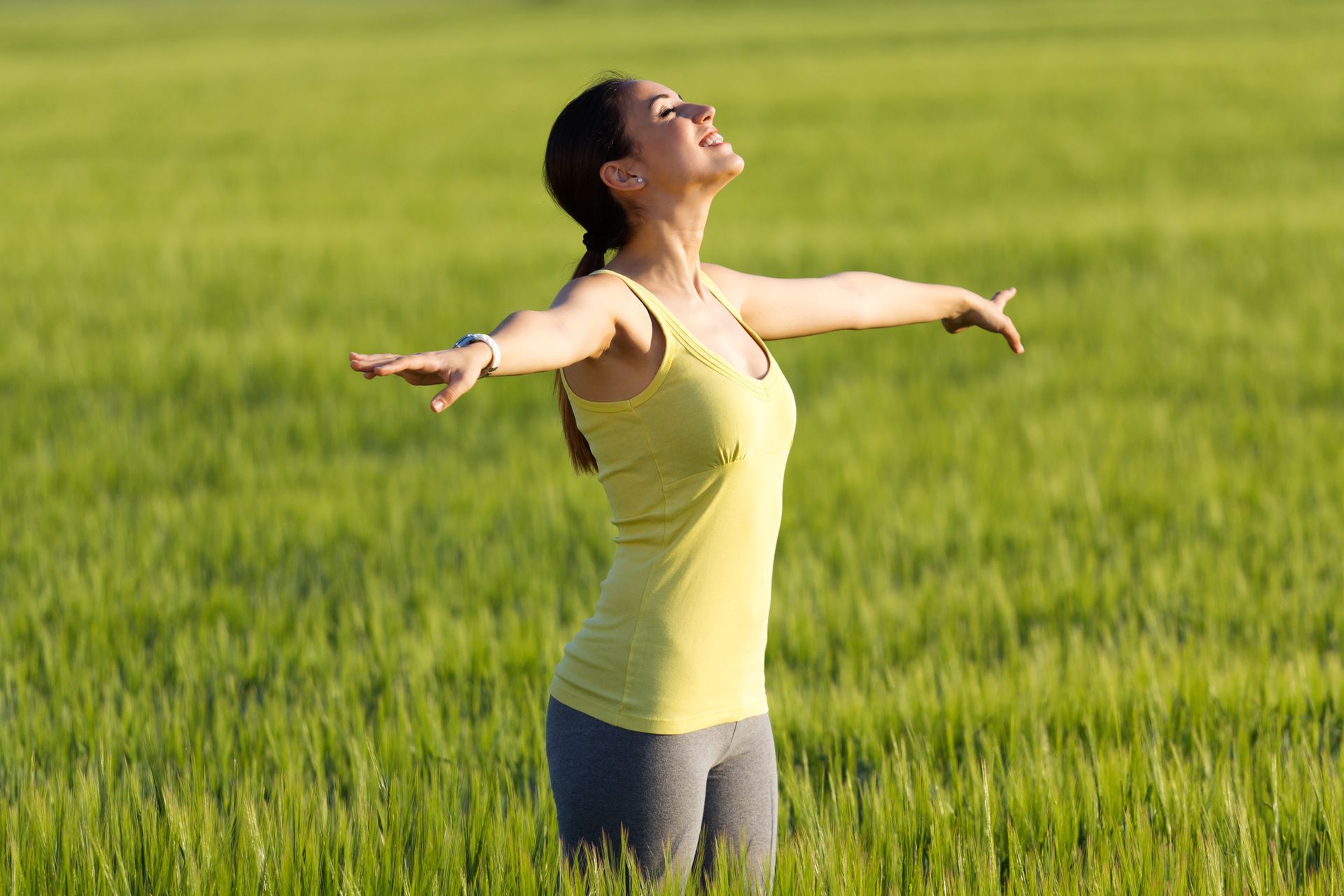 Woman standing with arms outstretched in a green field, facing upward in a yellow tank top