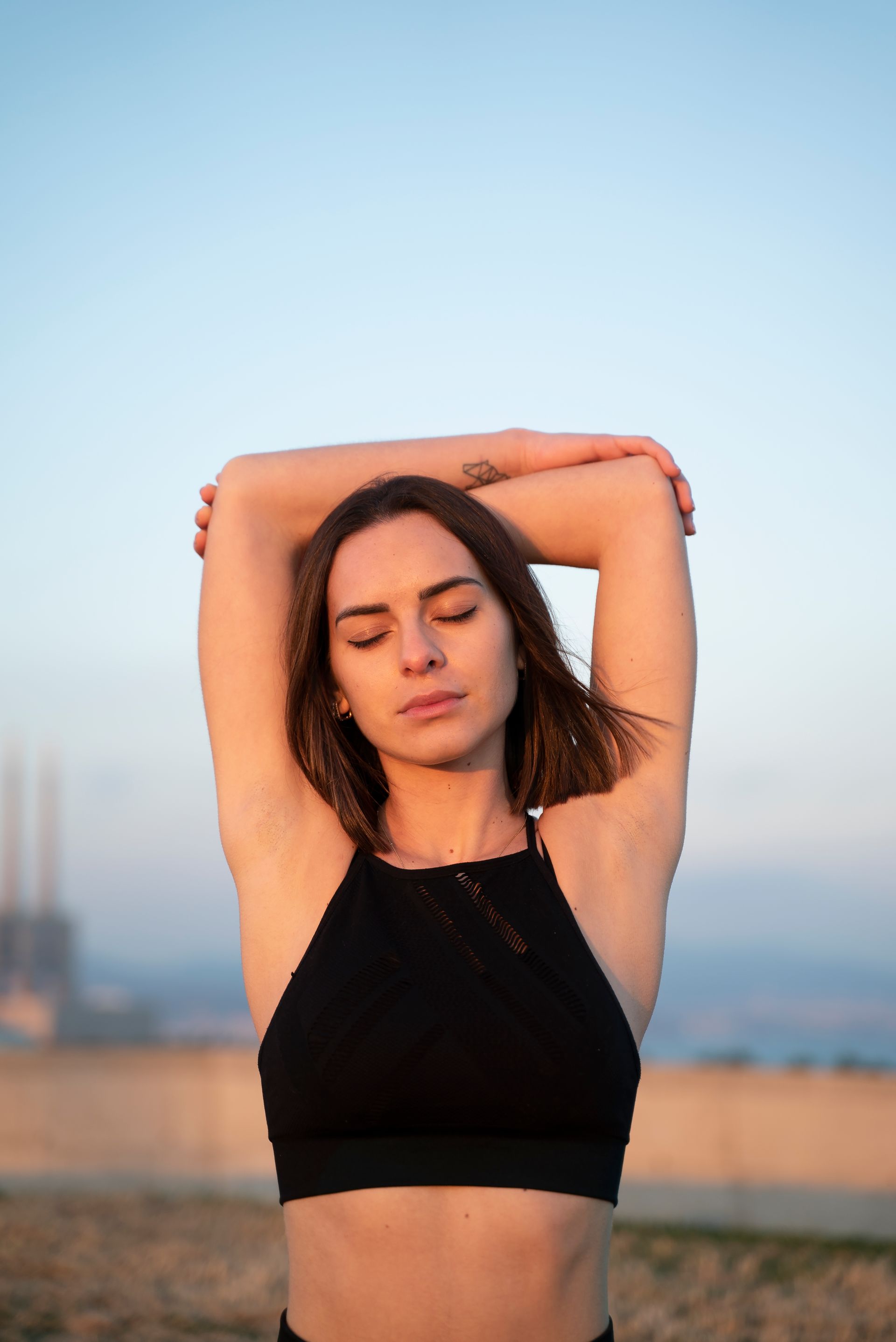 Woman in a black top stretching with arms raised outdoors at sunset, eyes closed.