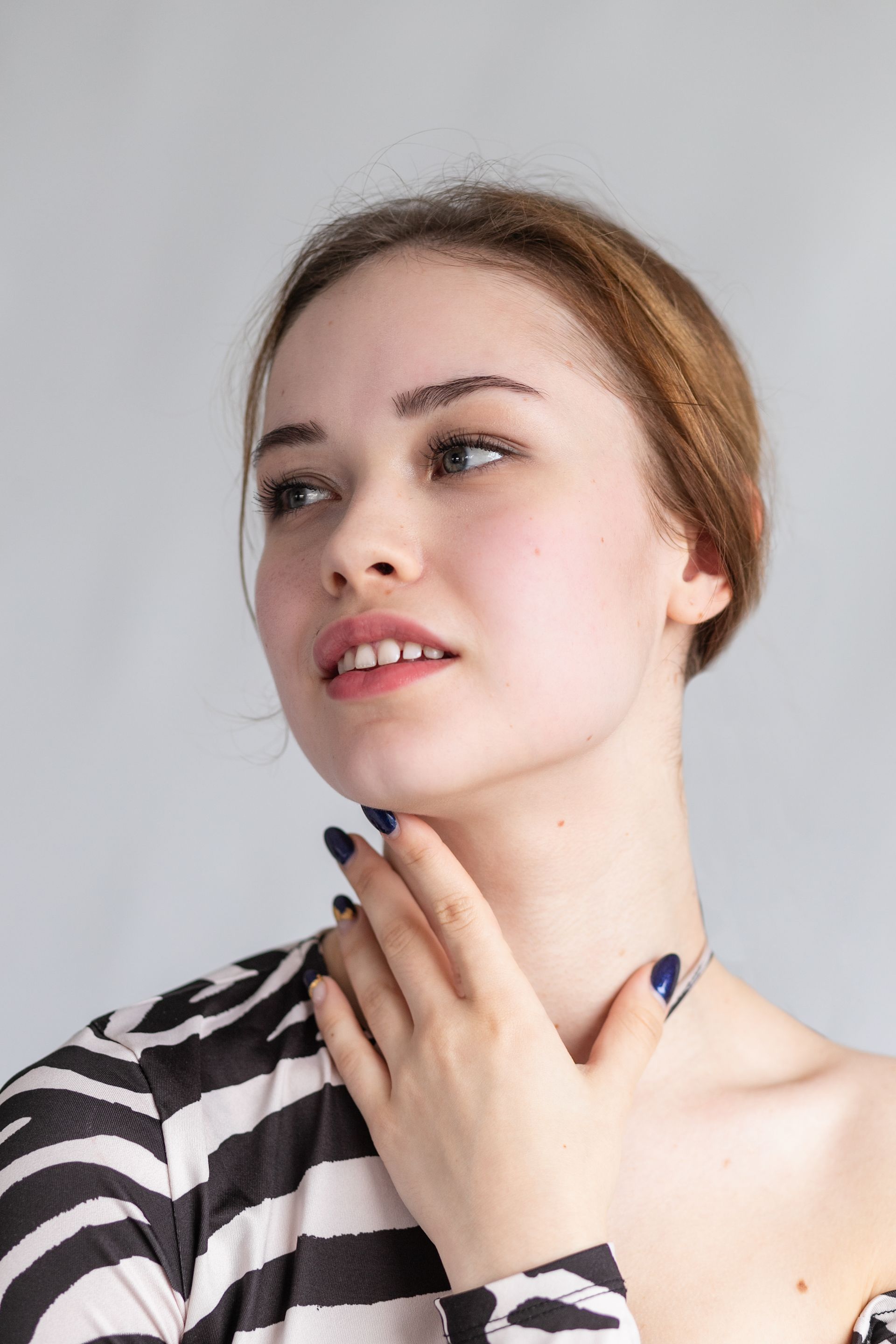 Portrait of a person in a black-and-white striped top touching their neck, looking upward against a light gray background