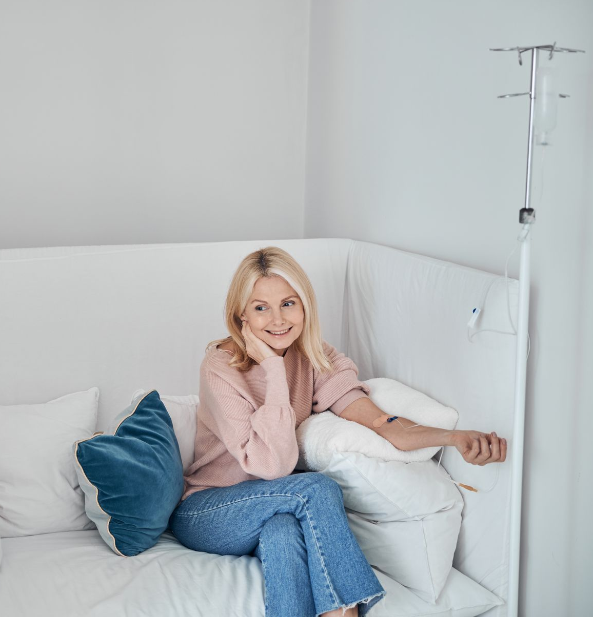 Person smiling on a white couch with blue and white pillows in a bright room, near an IV stand