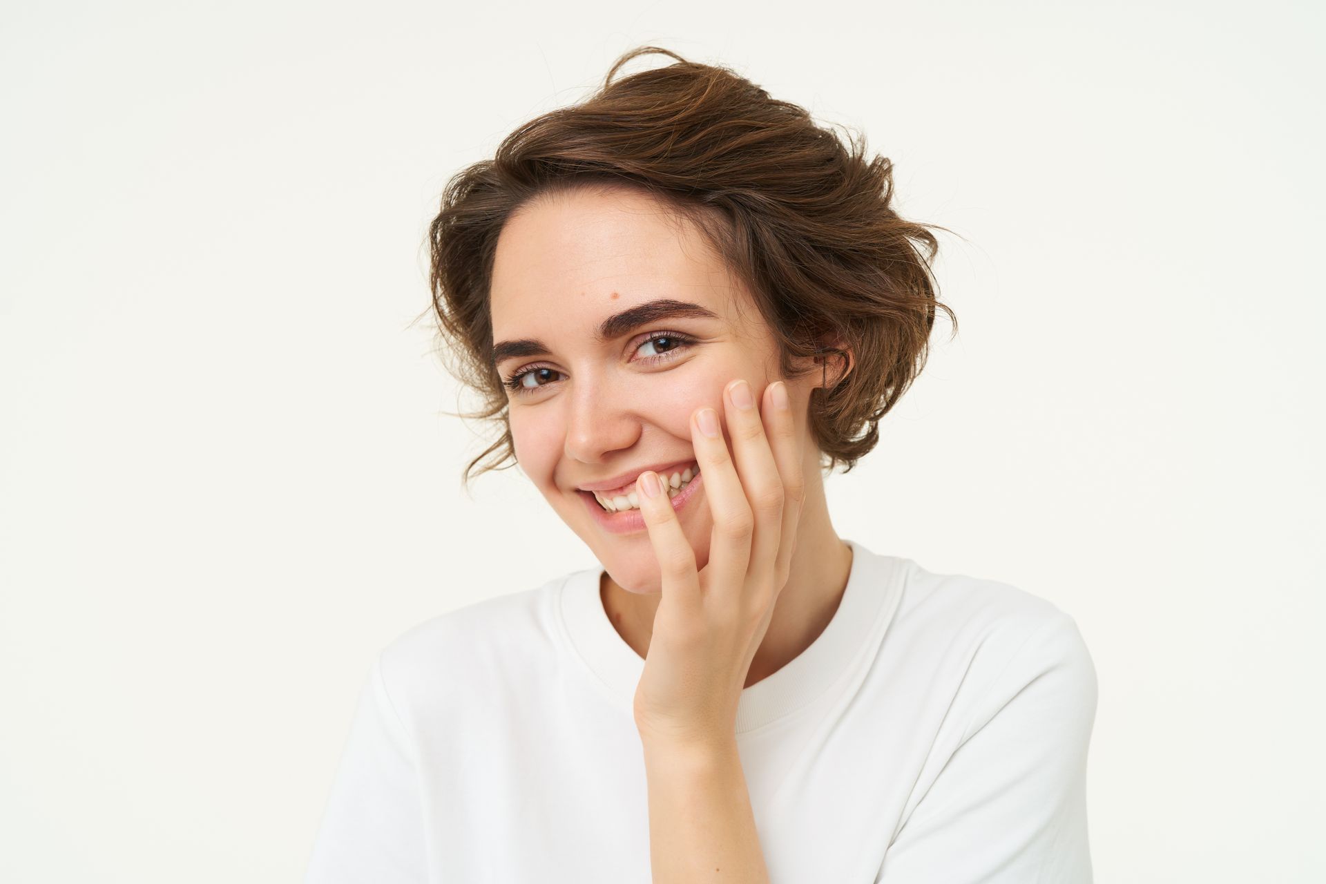 Smiling person with short brown hair, resting hand on cheek against a light background