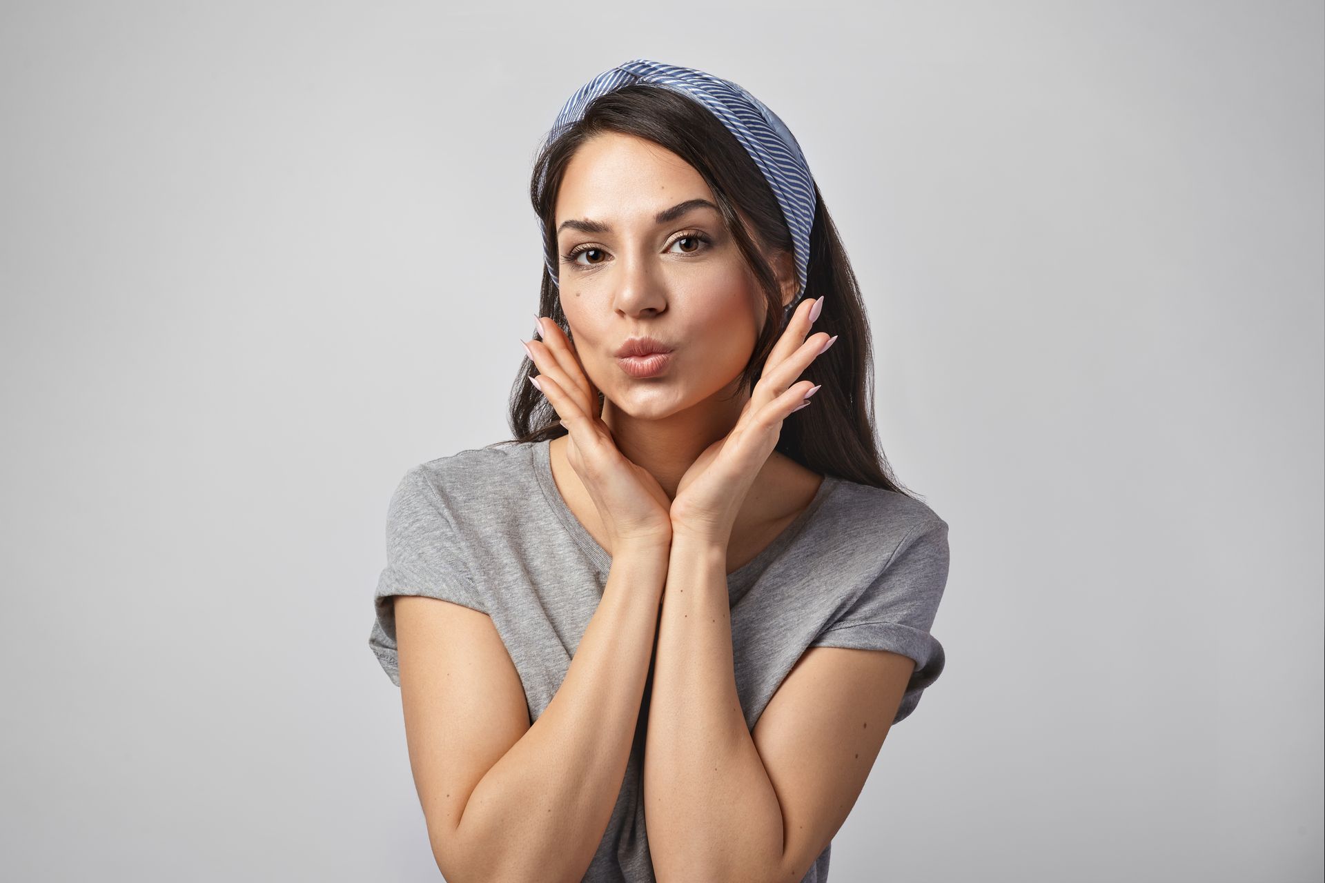 Woman in a gray shirt and blue headband posing with hands on cheeks against a plain background