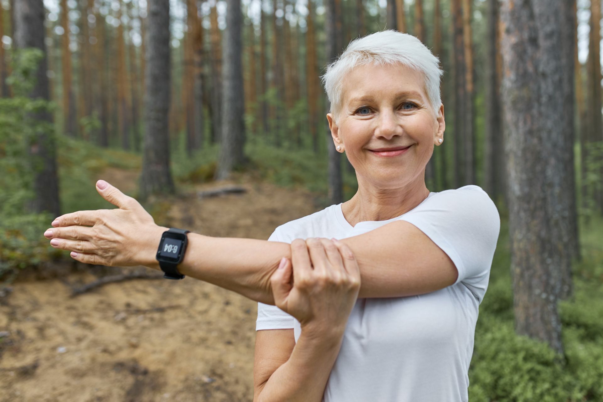 Woman stretching her arm outdoors in a forest, wearing a white shirt and black smartwatch.