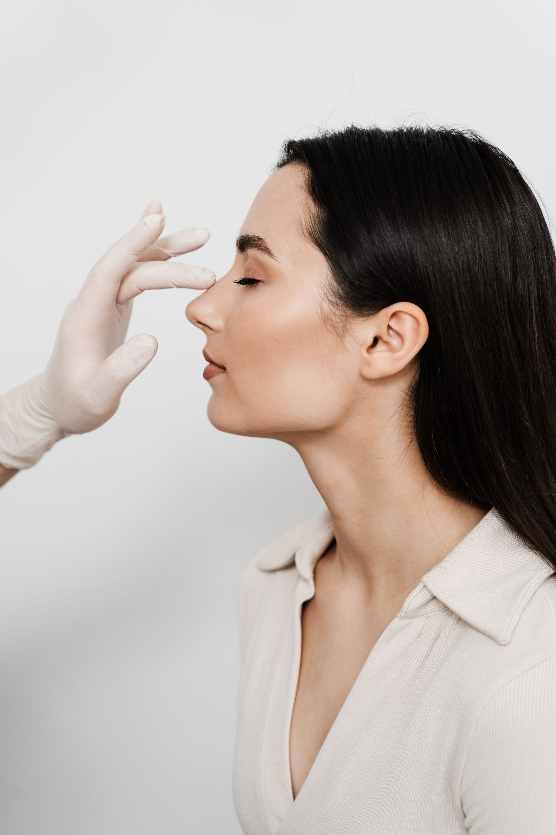 Person in profile as a gloved hand applies makeup or skincare to the nose on a white background