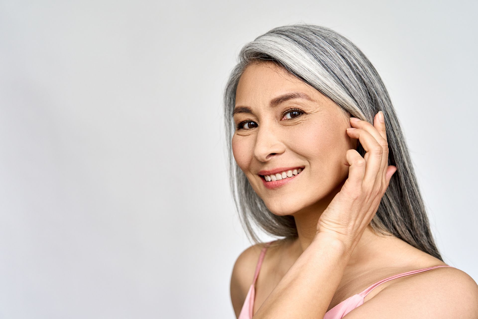 Smiling woman with silver hair touching her ear against a light gray background