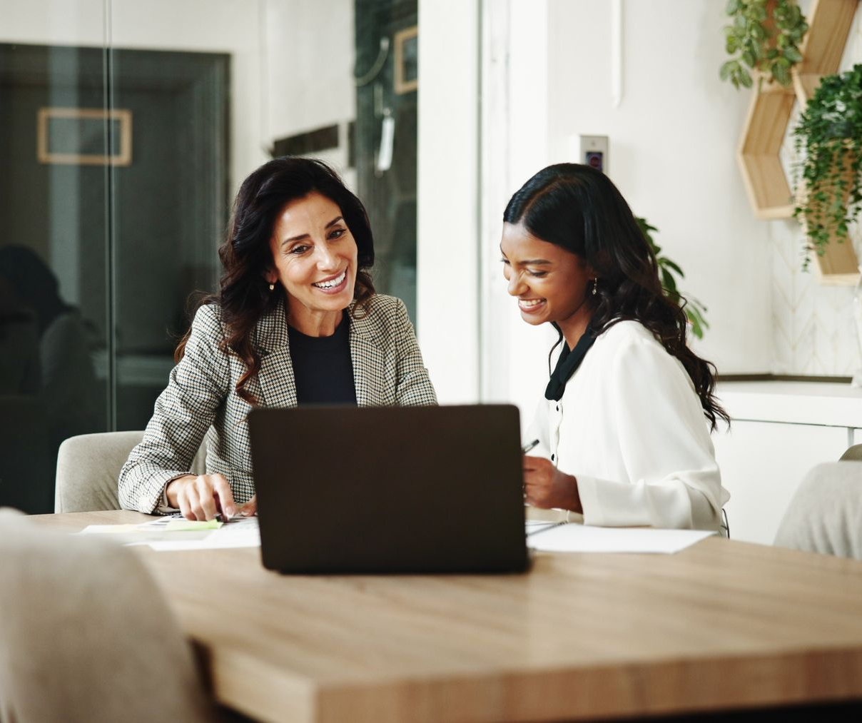 Two women smiling at a laptop across a wooden table in a bright office