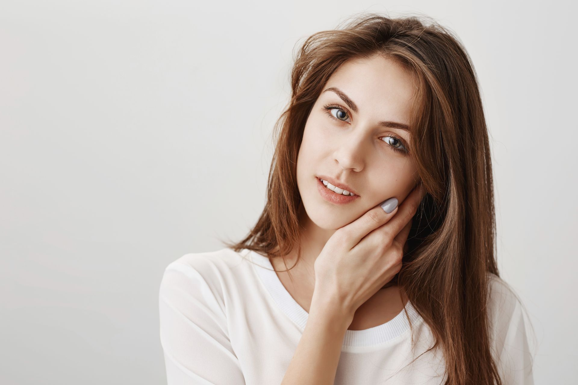 Woman in a white top touching her chin, smiling softly against a light gray background