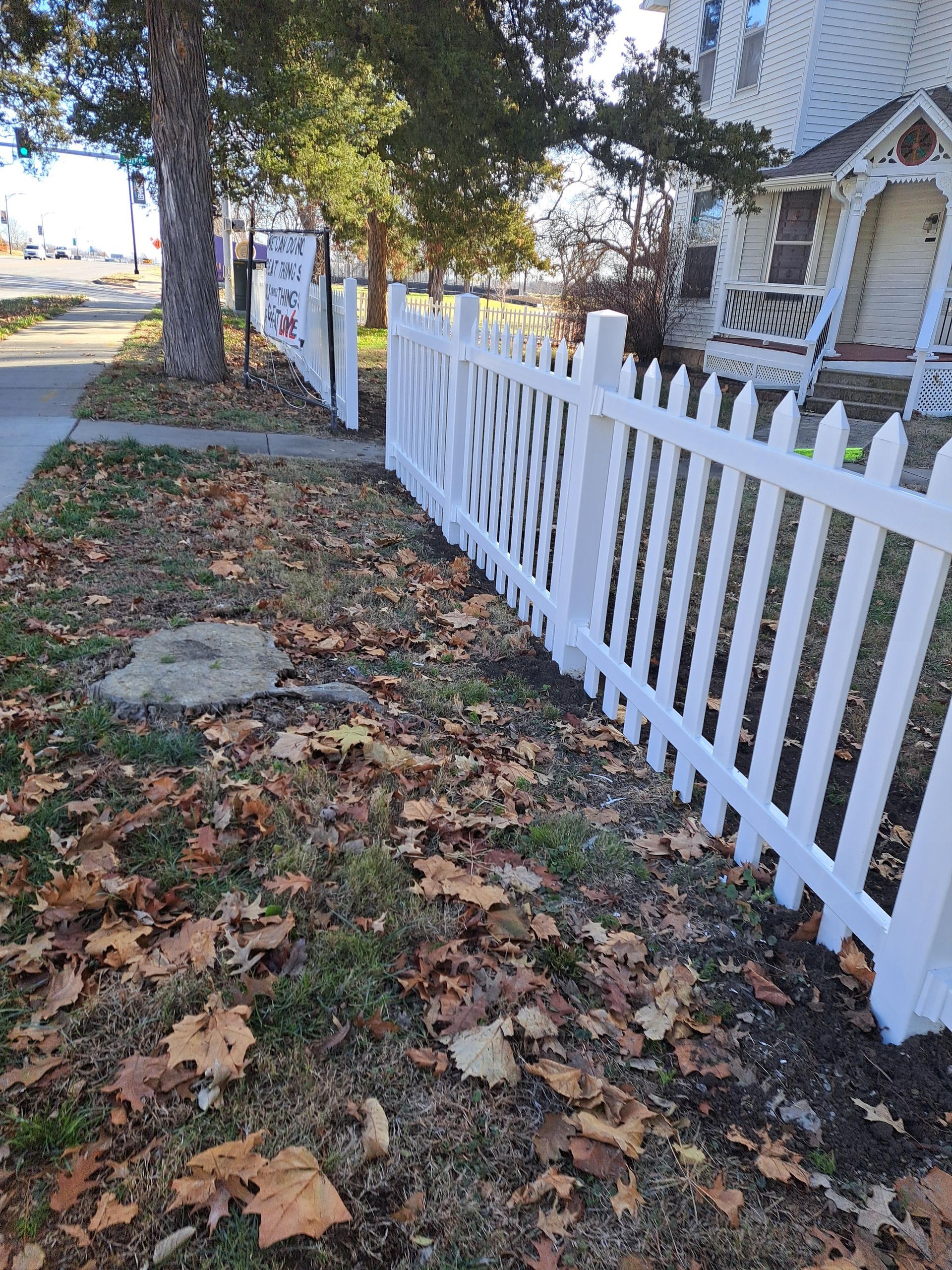 A white picket fence surrounds a yard with leaves on the ground.