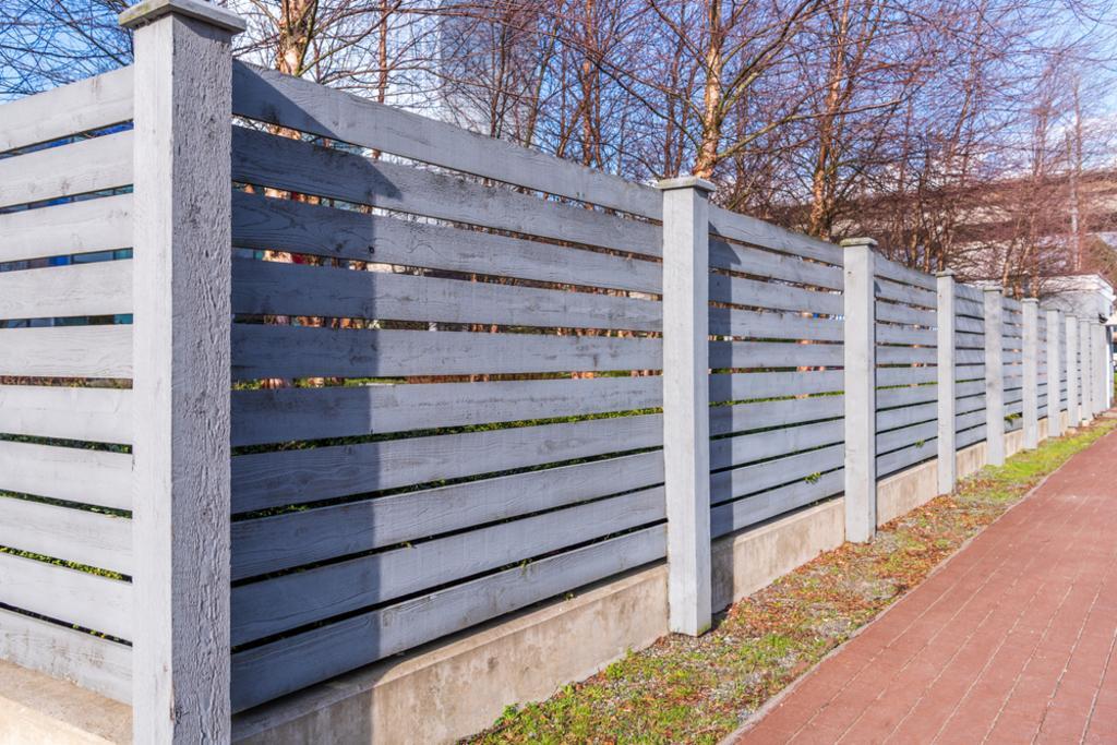 A wooden fence along a sidewalk with trees in the background.