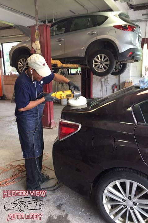 A worker polishes the trunk of a black car in an auto body shop, with another vehicle lifted on a hydraulic hoist above.