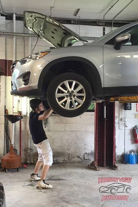 A person standing in an auto shop reaches up to touch a silver SUV suspended on a car lift.