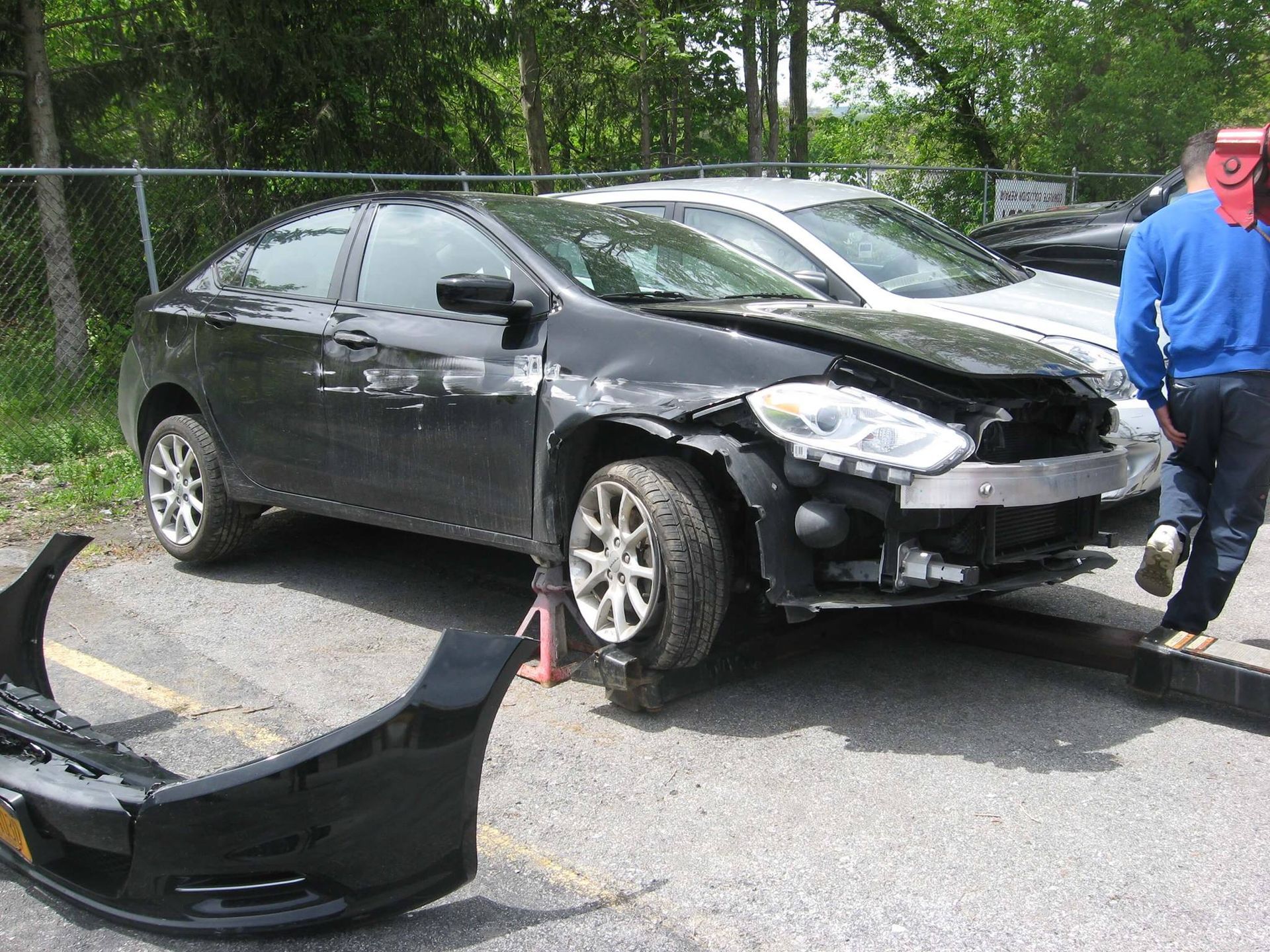 A damaged black car with its front bumper detached sits on a jack stand in a parking lot, with a person standing nearby.