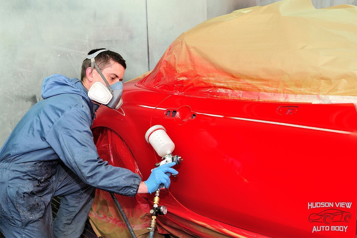 A person wearing protective gear and a respirator mask spray-painting a bright red car in a workshop.