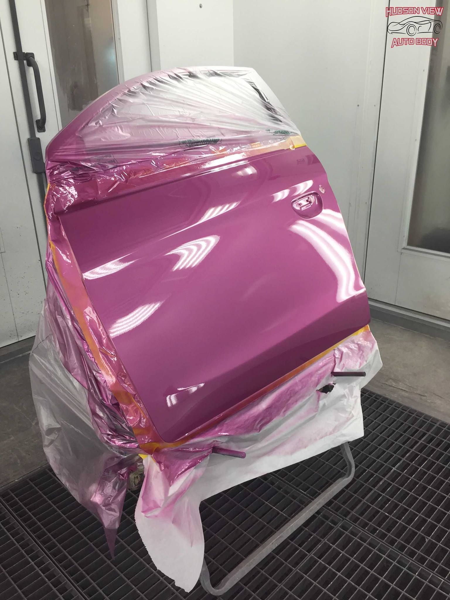 A freshly painted glossy magenta car door sits in a workshop spray booth, partially covered with plastic masking.