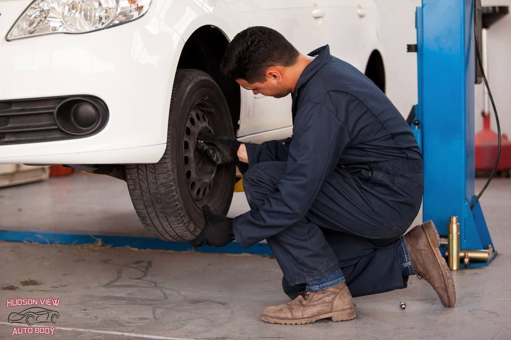 A mechanic in a navy jumpsuit kneeling to inspect or tighten a lug nut on a white car’s front tire in a garage.