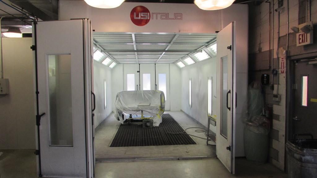 A white industrial paint booth with an open door and a plastic-covered vehicle centered inside.