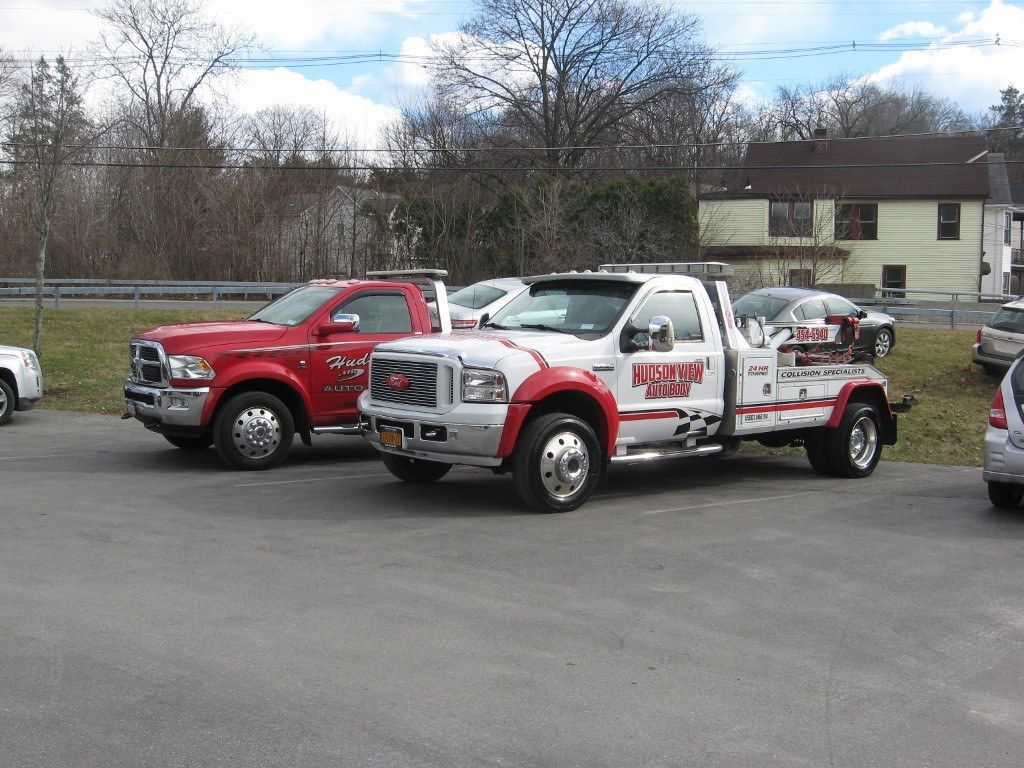 Two tow trucks, one red and one white, parked in an outdoor lot with trees and houses in the background.