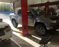 A grey pickup truck elevated on a hydraulic lift inside an auto repair shop, with a white car in the foreground.