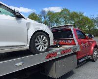 A white sedan with a dented passenger-side door is secured on the flatbed of a red Hudson View towing truck.