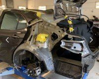 A damaged car in an auto body shop with its rear quarter panel and trunk interior exposed and undergoing repairs.