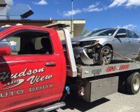 A silver sedan with front-end damage being transported on the flatbed of a bright red Hudson View Auto Body tow truck.