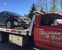 A damaged dark gray car secured on the flatbed of a red Hudson View Auto Body tow truck on a sunny day.