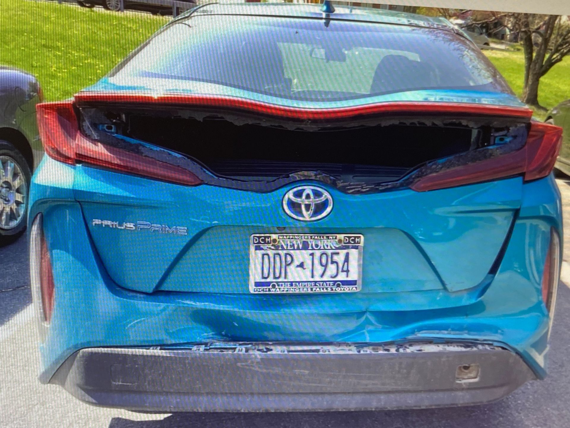 A damaged blue Toyota Prius Prime with a shattered rear window and a dented bumper parked outdoors.