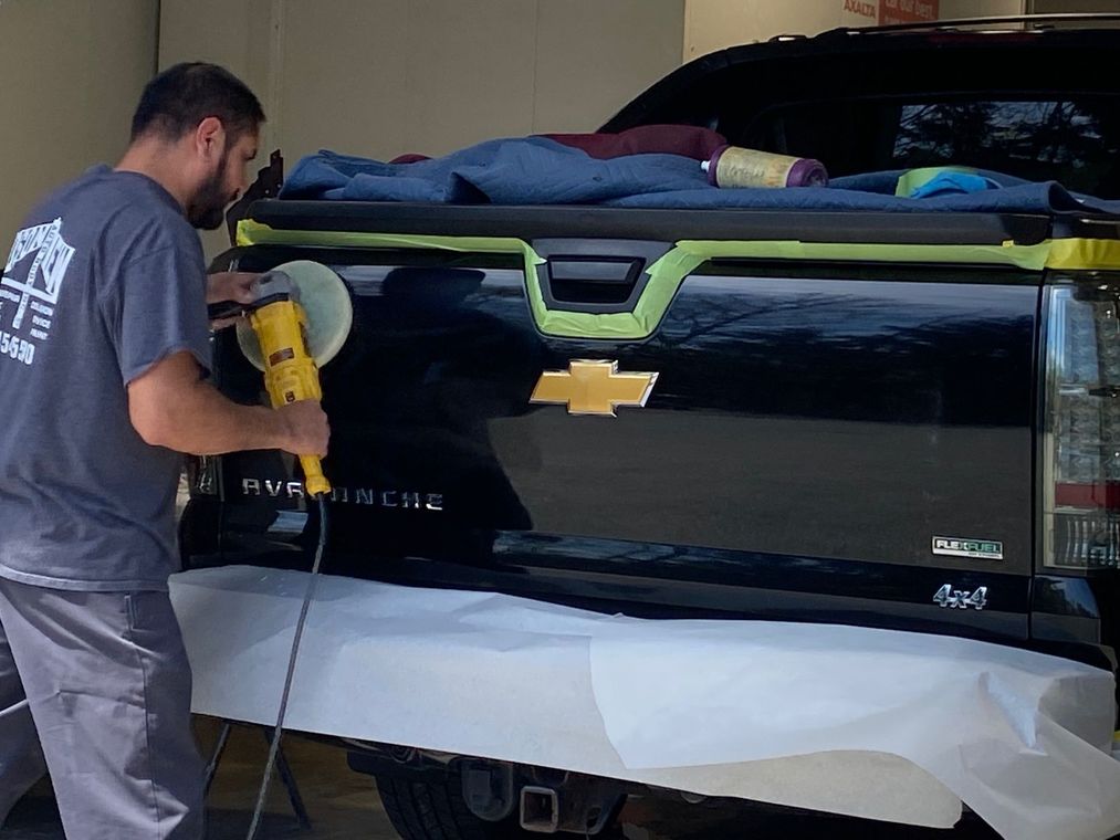 A worker uses a yellow power polisher to buff the black tailgate of a Chevrolet Avalanche truck protected by masking tape.