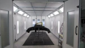 An automotive paint spray booth with a car bumper and door panel positioned on a stand in the center.